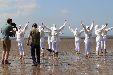 Eight dancers dressed in white stand barefoot in a circle facing away from the centre. They lift their arms and look up. The sand underfoot is pooled with water, which reflects the pale blue sky. A person operating a camera and another holding a boom film close to the group.