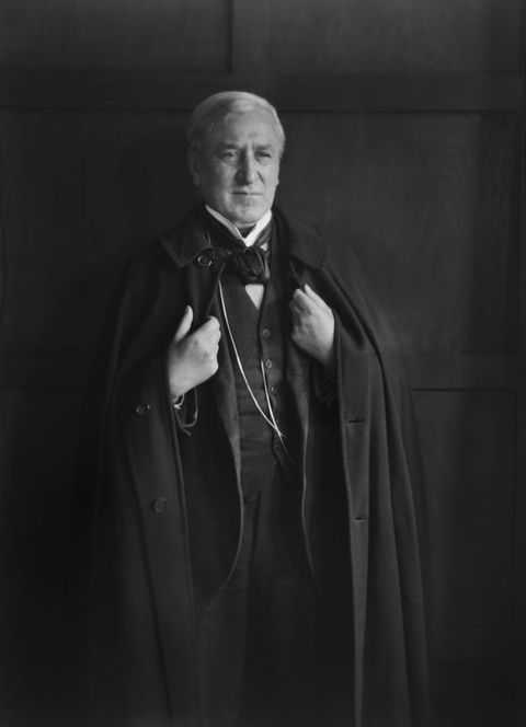 Black and white photograph. Shown from the knees up, a pale-skinned, clean shaven man stands looking off to our right from under knit brows. His white hair is combed over the top of his head, and wrinkles line his cheeks. Both hands grip the front edges of the long cape he wears over a three-piece suit. The wall behind him has dark panelling.