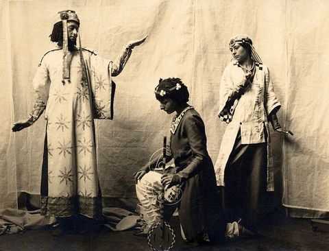 Black and white photograph. A man and two women pose against a draped background. They wear togas and headdresses. Two stand and one kneels, and most of their wrists are flexed so their hands are flat, palms up or down.