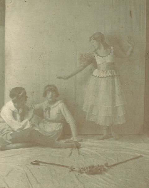 Black and white photograph. One woman in a white dress stands, arms outstretched as she looks at two people with brown hair, headbands, and bobs sitting to our left. The couple sit facing each other, legs wrapped around the other’s body.