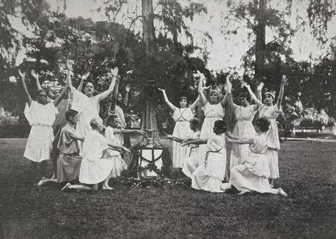 Black and white photograph. Seven men to our left and seven women to our right pose with arms raised or extended in a park or garden. They all wear white togas and are arranged around an altar-like table.