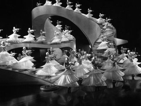 In black and white, at least three dozen women playing violins are arrayed across a staircase that twists up and around a film set. Their skirts each have three layers of hoops at different lengths, fanning out like nested trumpets’ horns to their feet.