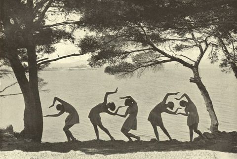 Black and white photograph. Five women wearing thigh-length togas pose in a line in the shadows between two trees against a body of water. Their arms and legs are posed at affected angles, resembling glyphs.