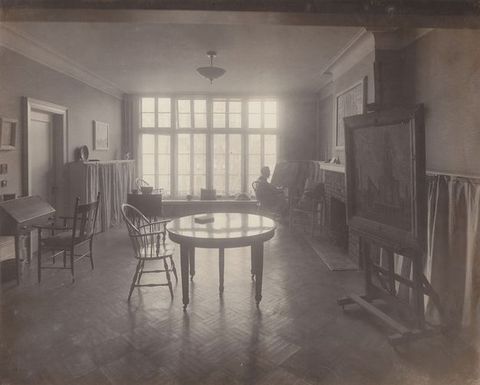 Black and white photograph. A round table and one chair is backlit by a wall of windows at the far end of this room. A desk, easel, and other furniture lines the walls. A sitting person is silhouetted near the windows.