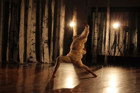 A dancer dressed in white sways back over widely planted feet, so his head disappears behind his torso. The front arm is raised. Strips of white fabric streaked with dark grey hang across the space behind the dancer, and he is illuminated by two spotlights glaring into the camera opposite us.