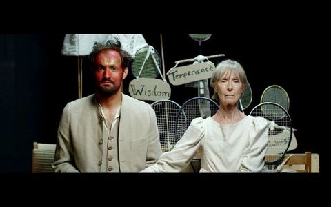 Shown from the waist up, a man with his face painted red and gold stands next to an older woman with grey bangs across her forehead. Her white dress has puffed cap sleeves, and the man wears a tan suit over an unbuttoned white shirt. Signs hanging from a stand made from tennis and badminton rackets behind them read “Wisdom” and “Temperance”.