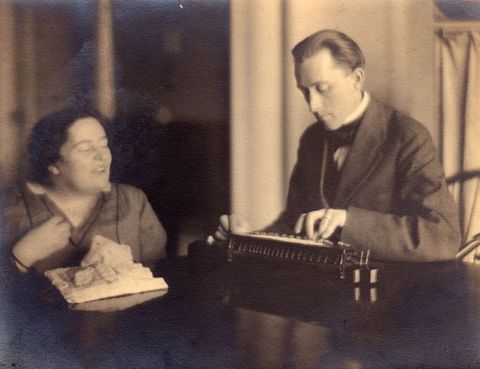 Black and white photograph. A woman and man sit at a high table. To our left, the woman holds one hand up to her chest and her eyes are closed as she seems to say something to the man. To our right, he looks down and touches a device, perhaps typing.