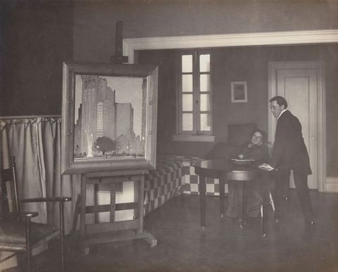 Black and white photograph. A painting of skyscrapers on an easel faces us, and two people sit and stand at a table farther back in the room to our right.