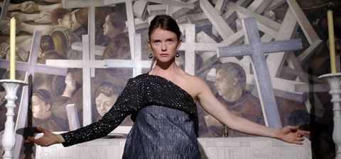 A pale-skinned woman holds her arms out wide in front of a painting showing a jumble of crosses and people. Her shimmering black shirt has one long sleeve and is off the other shoulder. A skirt in a shiny black material comes up under her bust.