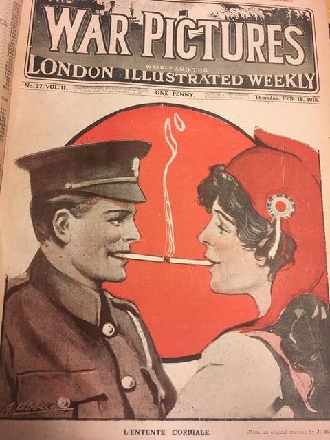 Cover of a magazine. Illustration of a man in uniform smoking a cigarette. He touches the burning ember to a cigarette smoked by a woman across from him. They both have pale skin and red-painted lips that smile around the cigarettes. The woman’s hat or scarf is also red, as is a disc behind the pair.