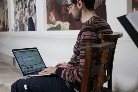 Colour photograph. A man sits in a wooden chair with a laptop open on his lap.