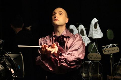 A bald, clean-shaven, pale-skinned man wears a billowing, rose-pink silk or satin shirt. He lifts his chin and looks intently out under raised brows. His hands are stretched out in front of him and one holds a conductor's baton. In the shadowy background, white hoods hangover signs reading “Wisdom” and “Temperance”.