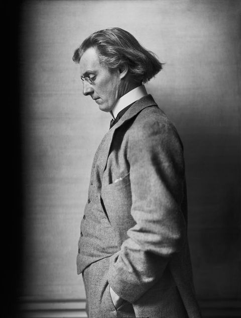 Black and white photograph. Shown from the hips up, a cleanshaven  man with chin-length hair and pale skin stands facing our left in profile. He looks down through pince-nez, and the hand we can see is in his pocket. His three-piece suit has a high white collar.