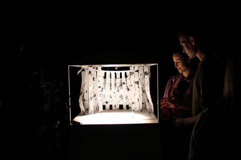 Colour photograph. Two people look at a model of a performance space, which has reflective material along the back wall and strips of paper or fabric hanging along the back and sides of the space. The paper is white with grey streaks and spatters. Three tiny white figures are on the model stage.