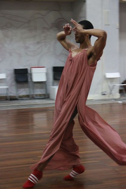 Colour photograph. A dancer steps forwards while leaning his torso back, creating a long diagonal. Both hands are raised with forefingers and thumbs pinched in front of his eyes. A swath of the rose-pink garment he wears flutters behind him.