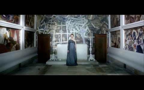 A woman wearing a black dress stands in a chapel, arms by her side. The wall behind her is painted with men struggling with and among a pile of white crosses.