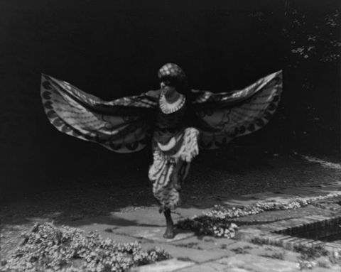 Black and white photograph. A dancer wearing a hawk costume bends one knee and raises that foot parallel to the ground. Both arms swoop out, and he looks off to our left through a black-eyed mask.