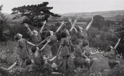 Black and white photograph. Twelve children wearing tunics pose in a field of wildflowers. They all face our right and pose with one or two arms raised.